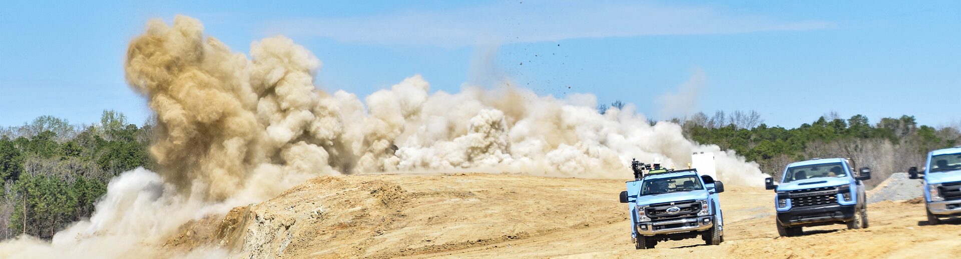A wide-format industrial banner showing a massive rock blasting explosion creating a large cloud of dust and debris on the left, with two heavy-duty off-road industrial pickup trucks and excavation equipment parked on-site against a clear blue sky.