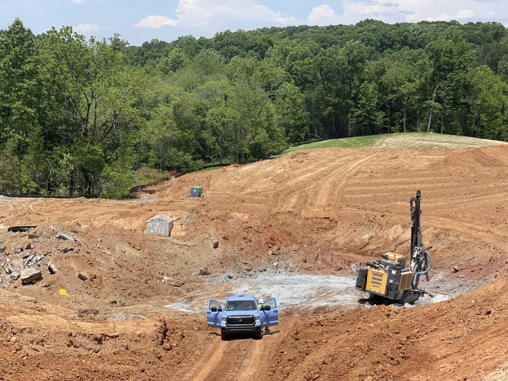 An overhead view of a massive civil construction site with a utility truck and heavy-duty rock drilling rig preparing the terrain.