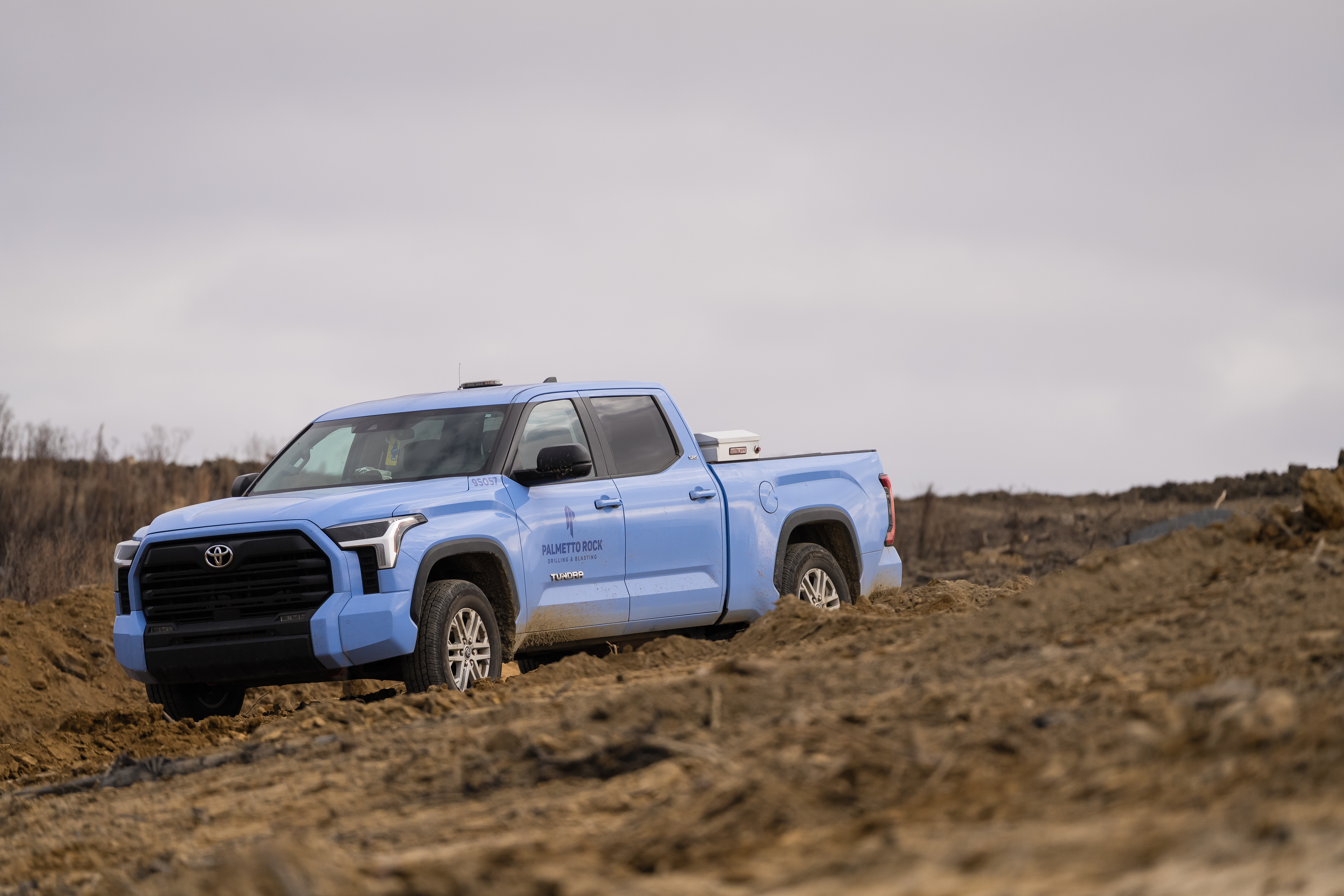 A light blue Palmetto Rock branded pickup truck parked on a muddy commercial site preparation and heavy civil construction excavation terrain.