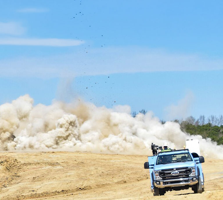 A blue truck speeds away from a massive dust cloud caused by an explosion on a sandy terrain, under a clear blue sky, conveying urgency and action.